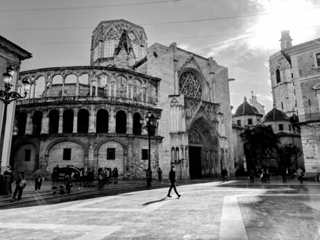 A monochrome, eye-level shot captures the Valencia Cathedral in Spain, bathed in bright sunlight. The cathedral's facade, with its intricate Gothic details, dominates the right side of the frame. A large rose window and pointed archways are prominent features. To the left, another historic building with arched windows and a curved balcony complements the cathedral's grandeur. Several people are seen walking across the paved square in front of the buildings, casting long shadows. The sky is bright, with the sun's glare visible towards the upper right corner.