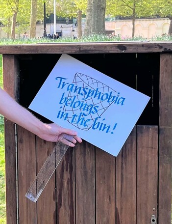 Homemade placard with image of a waste bin and blue text: Transphobia belongs in the bin. A hand is holding it in front of a wooden bin.