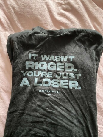 A black t-shirt with a pale pink gray background, the shirt has gray writing that reads “it wasn’t rigged. You’re just a loser. Meidas touch.”
