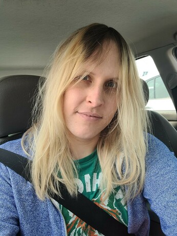 smiling trans girl with chaotic long bleache blonde hair with roots showing at the top. she's wearing a blue zipup