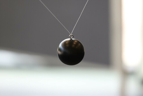 Close-up of a small metal sphere with an eyelet and a string running through the eyelet. The dark sphere is hanging mid-image against an unfocused bright background.