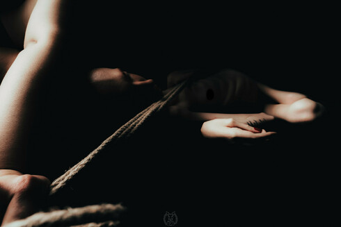 Ein Dunkel und sinnlich ausgeleuchtetes Foto von mir. Ich liege auf dem Rücken und bin nackt. Der Fokus liegt auf der Spannung des Seils, das eine Frau über meinen Körper spannt und dem Lichtspiel auf Haut, Arm und Hand. Die Szene wirkt intim, ruhig und selbstbestimmt. Eine Tätowierung und rot lackierte Fingernägel sind sichtbar – Zeichen von Individualität und Selbstinszenierung. Das Bild spielt mit Schatten und Körperlichkeit, ohne voyeuristisch zu wirken.