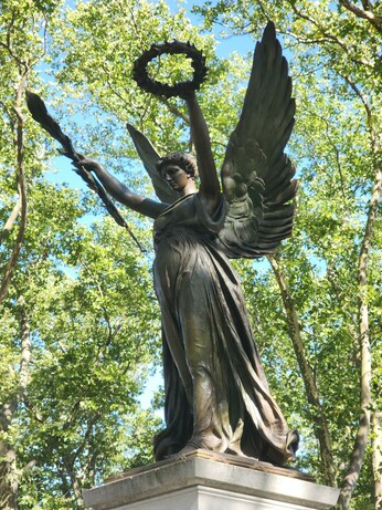 Bronze statue of an angel holding a palm frond on one hand and a laurel wreath in the other. It is surrounded by trees with a bit of blue sky peeking through the leaves. This is a recent photo of the Civil War memorial, not the postcard.