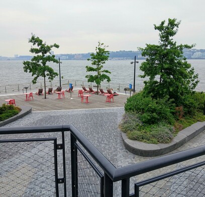 Photo on a gray day of a park on a pier, looking out toward the quiet Hudson River,  with scattered trees and a wooden area with orange chairs for lounging.