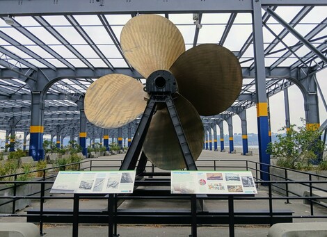 Very large copper-colored propeller, displayed in front of an open park structure converted from a tow building, on the Hudson river.