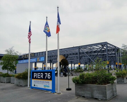Land entrance to Manhattan's Pier 76 park, with three flagpoles in front of a very large skeletal roof structure.
