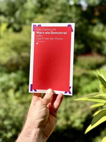 Eine linke Hand hält das beworbene Buch in die Kamera. Hintergrund: das Grün von Pflanzen. Das Buch trägt Titel den Titel: »Marx als Demokat oder: Das Ende der Politik«. Der Text steht auf einer roten Fläche, die an allen vier Ecken mit jeweils einem Buchstaben von MARX fixiert scheint, hinter der roten Fläche, d.h. die lila Buchstaben sind verdeckt.